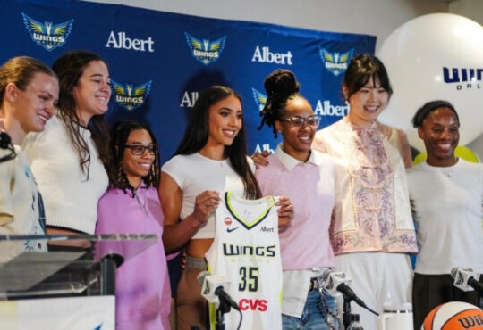 Azzi Fudd holds up her new #35 Dallas Wings jersey while posing with teammates Maddy Siegrist, JJ Quinerly, Grace Berger, Aziaha James, Li Yueru, and Shyanne Sellers at her introductory press conference.