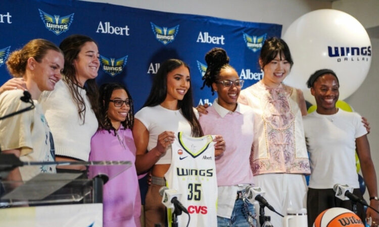 Azzi Fudd holds up her new #35 Dallas Wings jersey while posing with teammates Maddy Siegrist, JJ Quinerly, Grace Berger, Aziaha James, Li Yueru, and Shyanne Sellers at her introductory press conference.