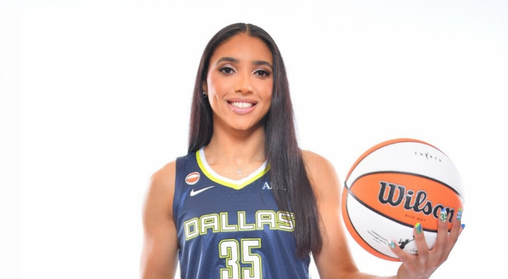 Azzi Fudd in a navy blue Dallas Wings Nike Explorer Edition jersey holding a basketball at her 2026 introductory press conference.