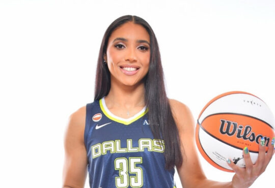 Azzi Fudd in a navy blue Dallas Wings Nike Explorer Edition jersey holding a basketball at her 2026 introductory press conference.