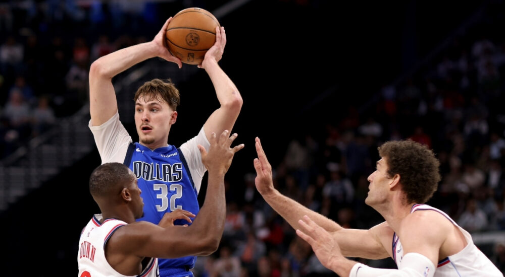 Cooper Flagg #32 of the Dallas Mavericks looks to pass while being guarded by Kris Dunn #8 and Brook Lopez #11 of the Los Angeles Clippers at Intuit Dome.