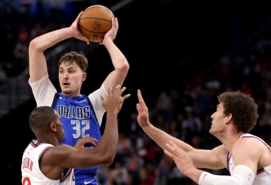 Cooper Flagg #32 of the Dallas Mavericks looks to pass while being guarded by Kris Dunn #8 and Brook Lopez #11 of the Los Angeles Clippers at Intuit Dome.