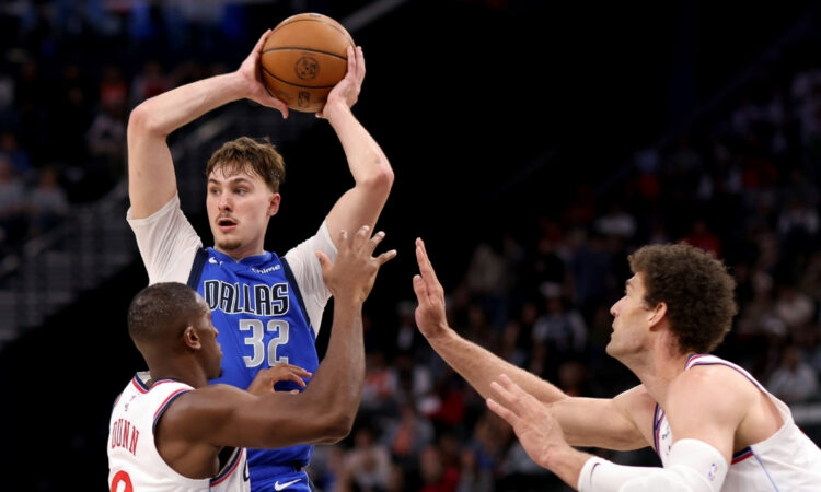 Cooper Flagg #32 of the Dallas Mavericks looks to pass while being guarded by Kris Dunn #8 and Brook Lopez #11 of the Los Angeles Clippers at Intuit Dome.