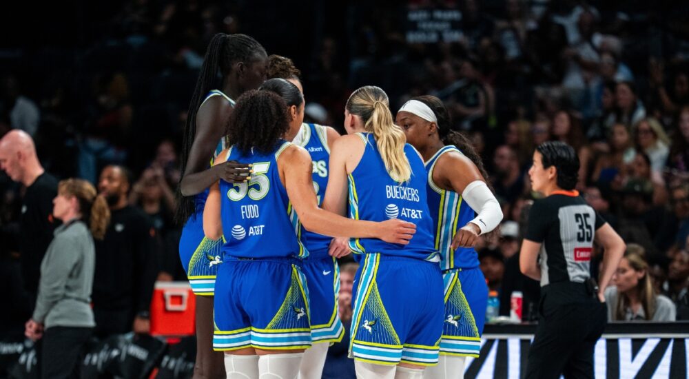 Dallas Wings players in blue road uniforms huddle together during a timeout against the Las Vegas Aces, with Azzi Fudd and Paige Bueckers visible from behind.