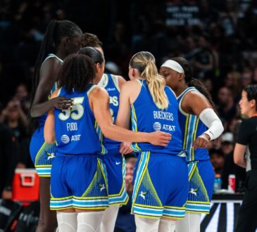 Dallas Wings players in blue road uniforms huddle together during a timeout against the Las Vegas Aces, with Azzi Fudd and Paige Bueckers visible from behind.
