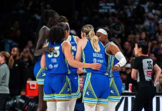 Dallas Wings players in blue road uniforms huddle together during a timeout against the Las Vegas Aces, with Azzi Fudd and Paige Bueckers visible from behind.