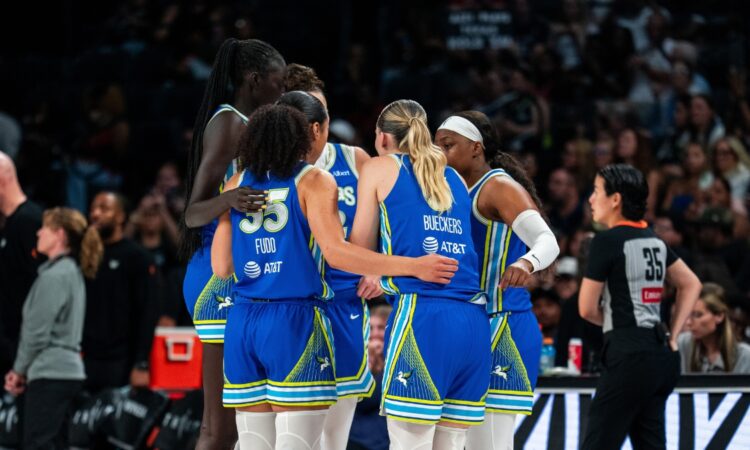 Dallas Wings players in blue road uniforms huddle together during a timeout against the Las Vegas Aces, with Azzi Fudd and Paige Bueckers visible from behind.