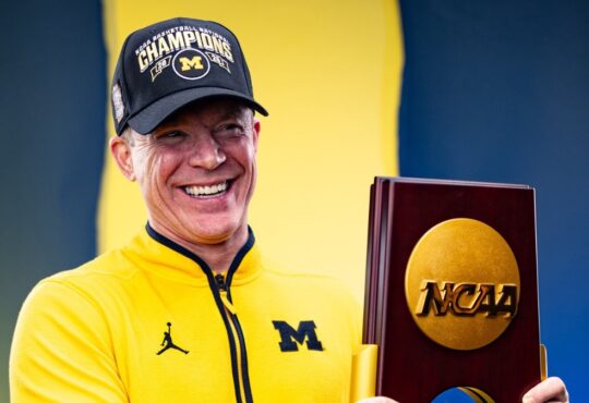 Dusty May smiles while holding the 2026 NCAA national championship trophy in a Michigan quarter-zip and Champions cap