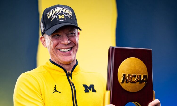 Dusty May smiles while holding the 2026 NCAA national championship trophy in a Michigan quarter-zip and Champions cap