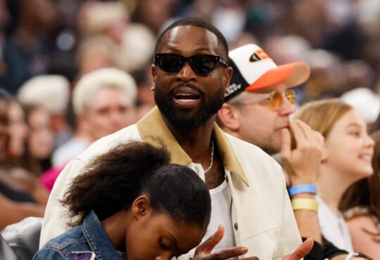 Dwyane Wade sits courtside in sunglasses and a cream jacket during the 2026 WNBA All-Star Game at United Center in Chicago.